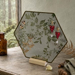 May include: A hexagonal jewelry display stand featuring a fine green mesh screen with a printed botanical leaf pattern. The screen is supported by a light-colored wooden base and holds several pairs of earrings, including gold-toned shapes and red heart-shaped dangles with white pearls.