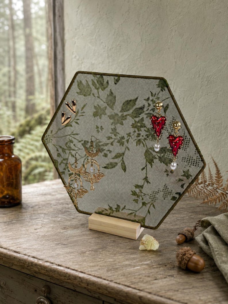May include: A hexagonal jewelry display stand featuring a fine green mesh screen with a printed botanical leaf pattern. The screen is supported by a light-colored wooden base and holds several pairs of earrings, including gold-toned shapes and red heart-shaped dangles with white pearls.