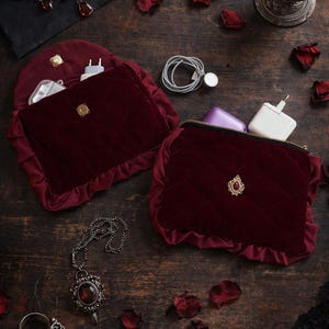 Könnte beinhalten: Zwei weinrote Samttaschen mit goldenen Akzenten und einem Steppmuster, zusammen mit einer passenden Halskette und einem Ring mit roten Edelsteinen. Ein Glas rote Flüssigkeit und verstreute Rosenblätter vervollständigen die Szene.