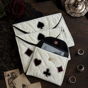 May include: Three white quilted fabric envelopes with black trim, each featuring rhinestone-encrusted playing card suit appliques. One envelope is open, revealing a black interior and a small electronic device with a visible screen.