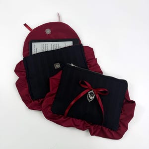 May include: Black and burgundy ebook reader case and matching zippered pouch. The case has a burgundy satin ruffle and a silver clasp. The pouch has a burgundy ribbon and a silver charm. The ebook reader is visible inside the case.