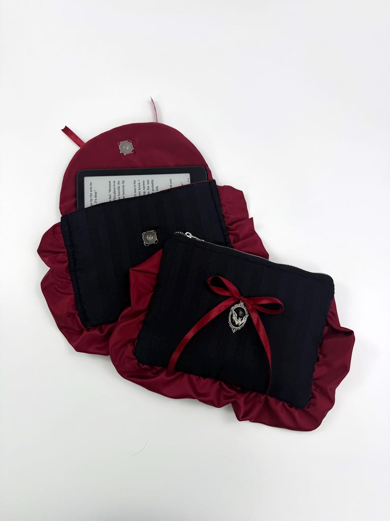 May include: Black and burgundy ebook reader case and matching zippered pouch. The case has a burgundy satin ruffle and a silver clasp. The pouch has a burgundy ribbon and a silver charm. The ebook reader is visible inside the case.