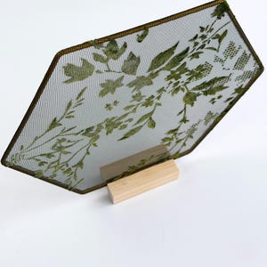 May include: A hexagonal metal mesh screen featuring a green botanical leaf pattern, framed in a thin dark metal border. The screen is supported by a rectangular light-colored wooden base, providing a stable upright display for the piece.