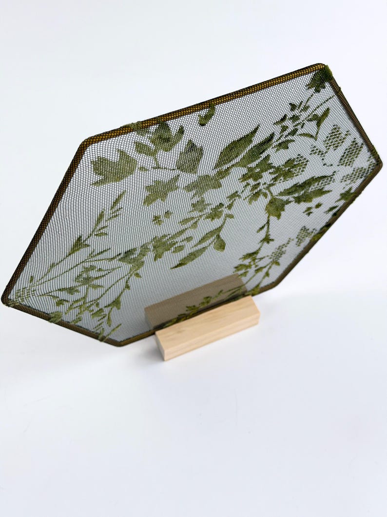 May include: A hexagonal metal mesh screen featuring a green botanical leaf pattern, framed in a thin dark metal border. The screen is supported by a rectangular light-colored wooden base, providing a stable upright display for the piece.