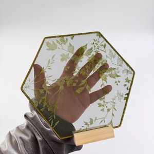 May include: A hexagonal piece of fine dark mesh fabric featuring a printed green botanical pattern of leaves and stems. The mesh is framed by a thin dark border and is supported at the base by a small, rectangular, light-colored wooden stand.