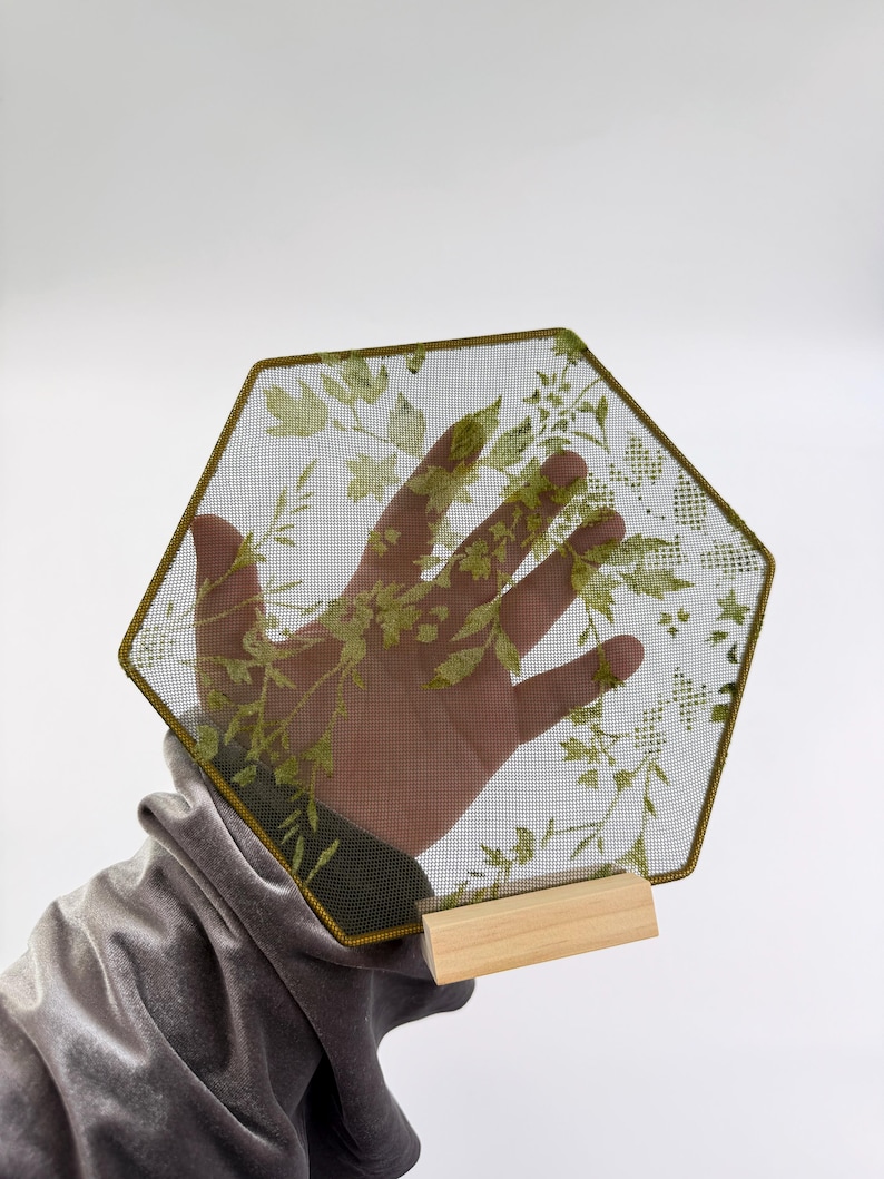 May include: A hexagonal piece of fine dark mesh fabric featuring a printed green botanical pattern of leaves and stems. The mesh is framed by a thin dark border and is supported at the base by a small, rectangular, light-colored wooden stand.