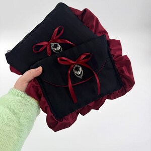 May include: Two black pouches with burgundy ruffled edges. Each pouch has a burgundy ribbon tied in a bow, with a silver-colored charm. The pouches are held together, showcasing the contrasting colors and textures.