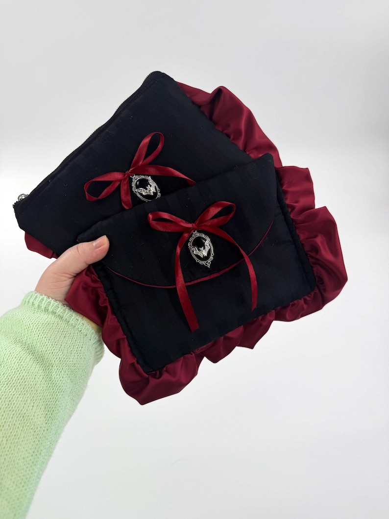 May include: Two black pouches with burgundy ruffled edges. Each pouch has a burgundy ribbon tied in a bow, with a silver-colored charm. The pouches are held together, showcasing the contrasting colors and textures.