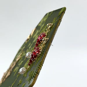 May include: A pair of gold-tone earrings featuring a dangling red chili pepper charm accented with a small white pearl at the base. The earrings are attached to a dark green textured card with a subtle gold floral pattern printed on the surface.