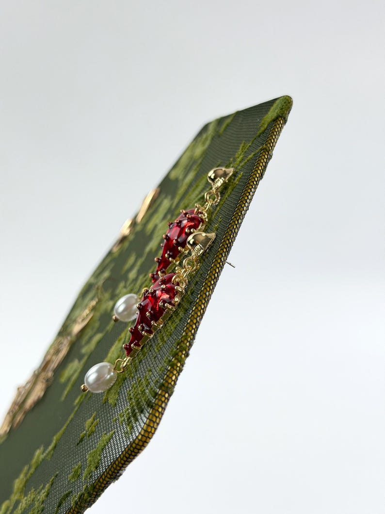 May include: A pair of gold-tone earrings featuring a dangling red chili pepper charm accented with a small white pearl at the base. The earrings are attached to a dark green textured card with a subtle gold floral pattern printed on the surface.