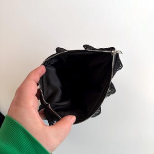 May include: Black lace pouch with a silver zipper, open to reveal a black interior. The pouch has a delicate, scalloped lace trim around the edges. The background is plain white.