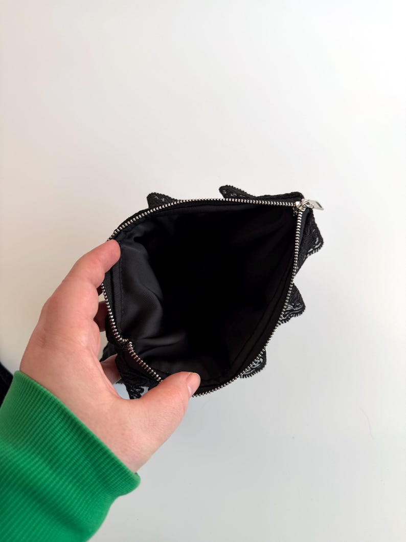 May include: Black lace pouch with a silver zipper, open to reveal a black interior. The pouch has a delicate, scalloped lace trim around the edges. The background is plain white.