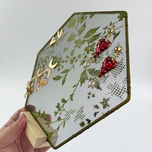 May include: A hexagonal jewelry display stand featuring a green mesh screen with a printed botanical leaf pattern. The stand is supported by a light-colored wooden base and holds several gold-toned earrings, including red heart-shaped drops with white pearls.