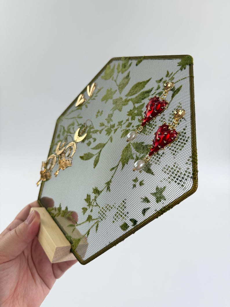May include: A hexagonal jewelry display stand featuring a green mesh screen with a printed botanical leaf pattern. The stand is supported by a light-colored wooden base and holds several gold-toned earrings, including red heart-shaped drops with white pearls.