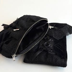 May include: Black velvet bag with an open zipper, revealing a dark interior. The bag features intricate lace detailing, satin ribbon accents, and a silver zipper. The design includes a matching black lace-trimmed accessory.