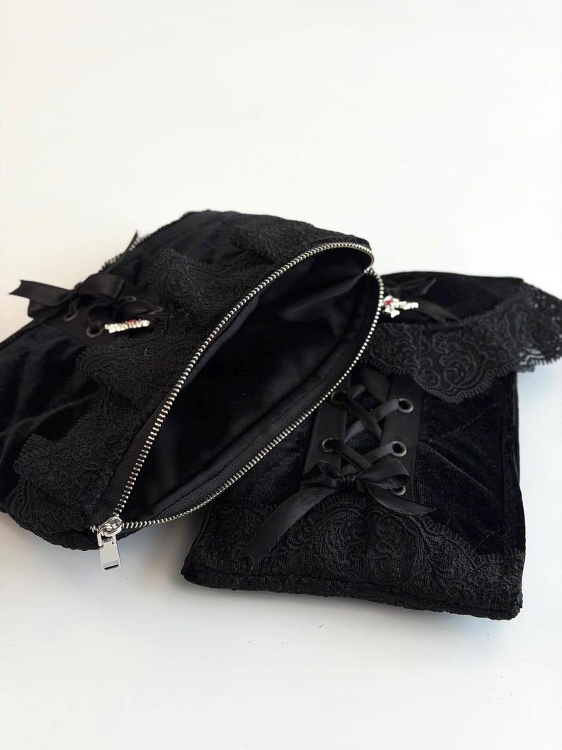 May include: Black velvet bag with an open zipper, revealing a dark interior. The bag features intricate lace detailing, satin ribbon accents, and a silver zipper. The design includes a matching black lace-trimmed accessory.