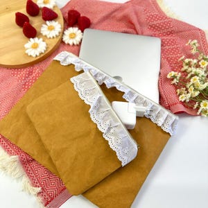 Könnte beinhalten: Zwei braune Beutel mit weißer Spitzenborte und Reißverschlüssen werden gezeigt. Ein Laptop und ein Ladegerät sind sichtbar. Die Beutel liegen auf einem roten Stoff mit Muster und weißem Fransen. Dekorative Erdbeeren und Gänseblümchen liegen auf einem Holzbrett.