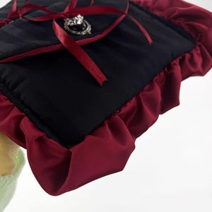 May include: Black and burgundy clutch with a striped pattern. The clutch features a burgundy ruffle trim and a burgundy ribbon tied in a bow. A silver-toned brooch with a black center stone adorns the front.