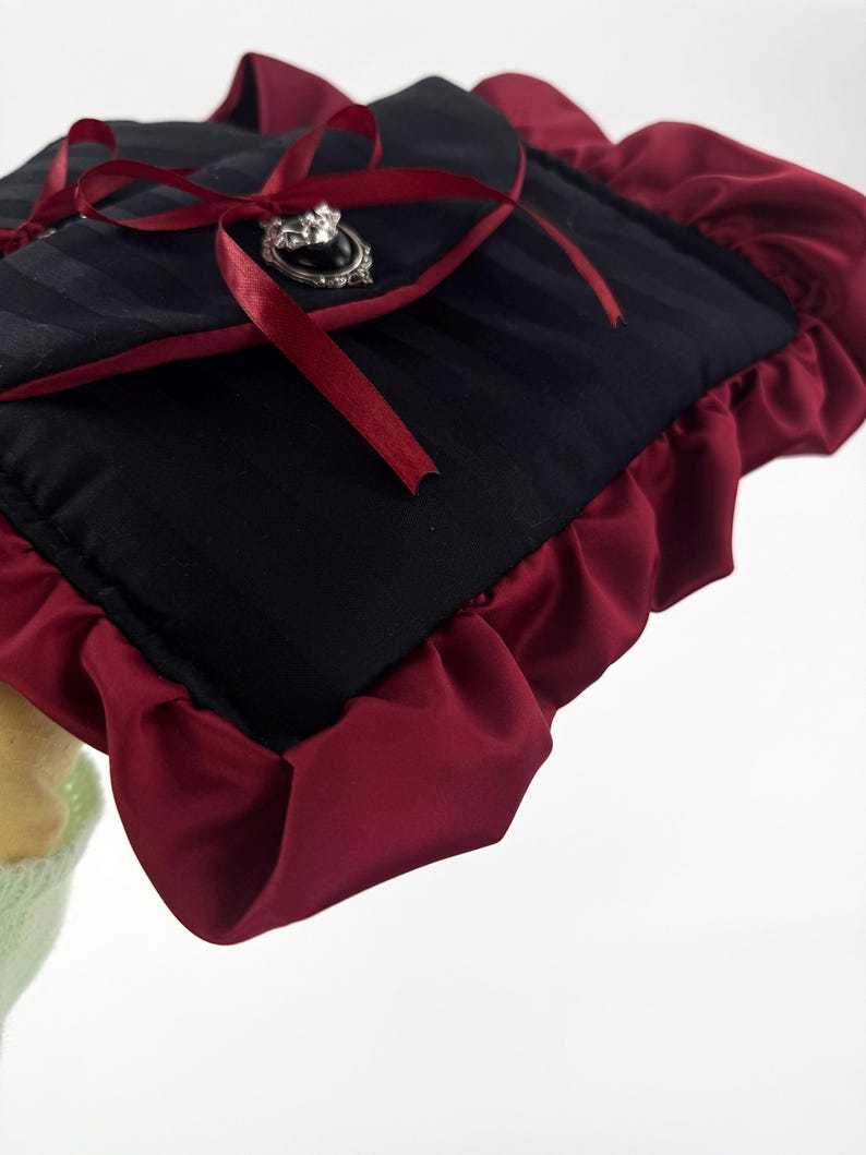 May include: Black and burgundy clutch with a striped pattern. The clutch features a burgundy ruffle trim and a burgundy ribbon tied in a bow. A silver-toned brooch with a black center stone adorns the front.