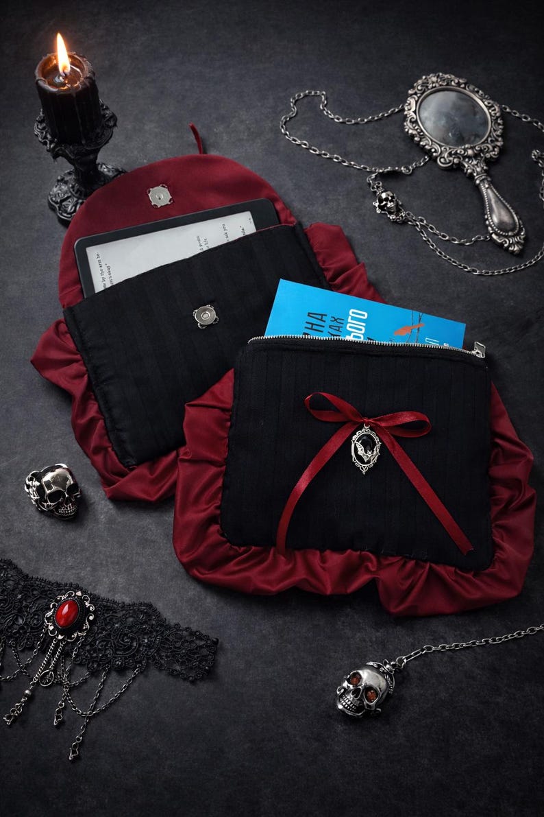 May include: Black and burgundy gothic-style pouches with a decorative bow and a silver pendant. One pouch is open, revealing a tablet and a book. Other gothic accessories include a skull ring, a hand mirror, and a black lace choker.