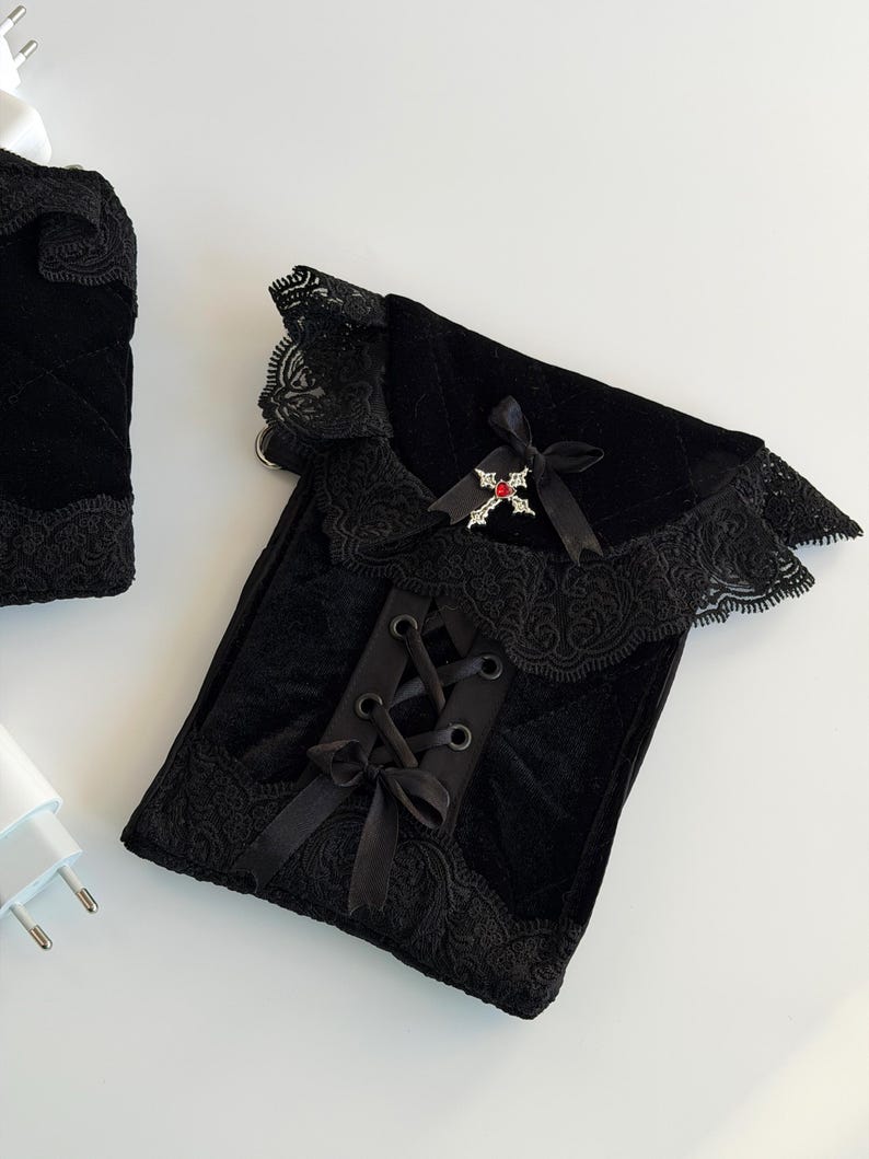 May include: Black velvet pouch with lace trim and a ribbon tie closure. The pouch features a decorative cross with a red gem and a black bow. The design is gothic-inspired.