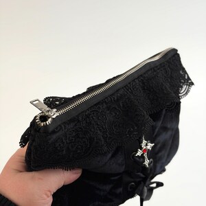 May include: Black velvet clutch with a silver zipper and lace trim. A silver cross with a red gem is attached to the front. The clutch has a black ribbon tie and is held in a person's hand.
