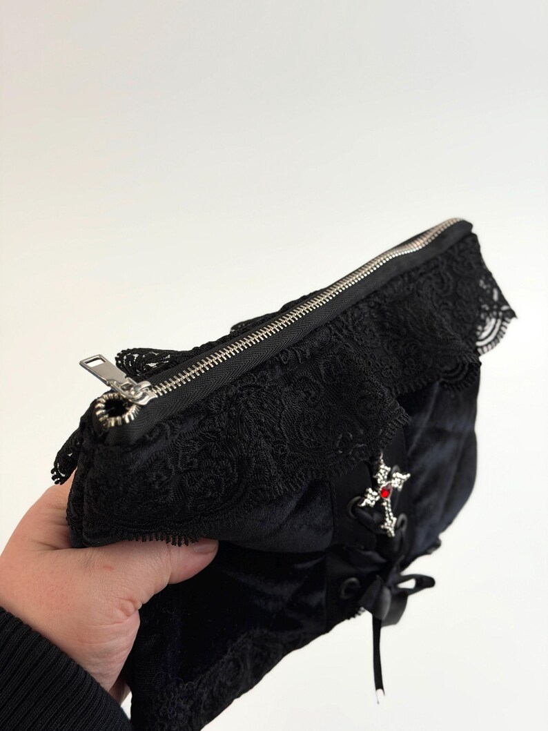 May include: Black velvet clutch with a silver zipper and lace trim. A silver cross with a red gem is attached to the front. The clutch has a black ribbon tie and is held in a person's hand.