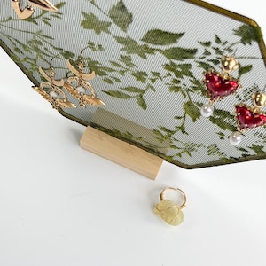 May include: A hexagonal jewelry display stand with a green botanical mesh screen and a light wood base. The stand holds gold-tone heart earrings, crescent moon earrings with white stones, and red heart earrings with pearls. A raw yellow stone ring sits below.