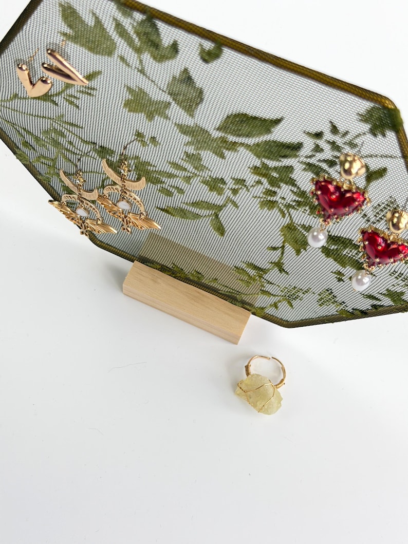 May include: A hexagonal jewelry display stand with a green botanical mesh screen and a light wood base. The stand holds gold-tone heart earrings, crescent moon earrings with white stones, and red heart earrings with pearls. A raw yellow stone ring sits below.