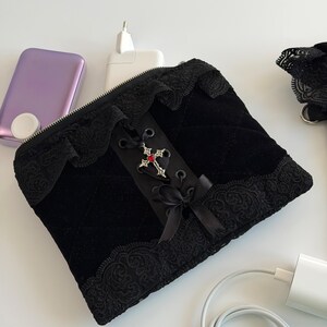 May include: Black velvet pouch with a decorative cross and lace trim. The pouch has a zipper closure and a ribbon bow. A purple device, a white charger, and a white cable are also visible.