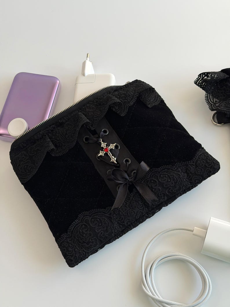 May include: Black velvet pouch with a decorative cross and lace trim. The pouch has a zipper closure and a ribbon bow. A purple device, a white charger, and a white cable are also visible.