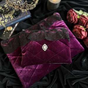 Op de afbeelding: Twee gewatteerde fluwelen pouches in diep paars, afgezet met zwarte kant. De kleinere pouch heeft een camee broche. Ze liggen op zwart fluweel, met een kroon, kaars en rozen op de achtergrond.