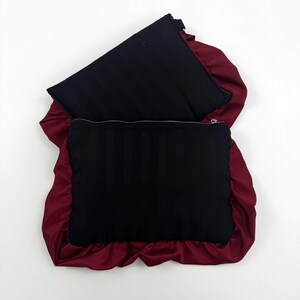 May include: Two black rectangular zippered pouches with vertical stripes, set against a deep red satin background. The satin creates a ruffled border around the pouches, adding a textural contrast.