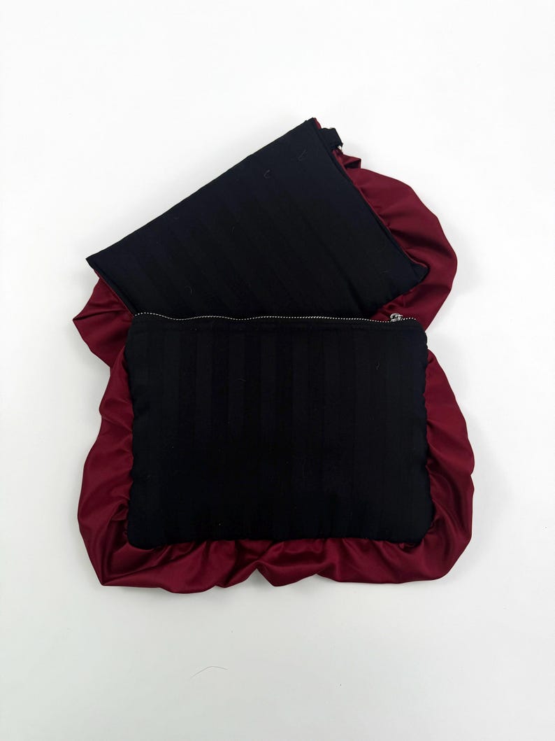 May include: Two black rectangular zippered pouches with vertical stripes, set against a deep red satin background. The satin creates a ruffled border around the pouches, adding a textural contrast.