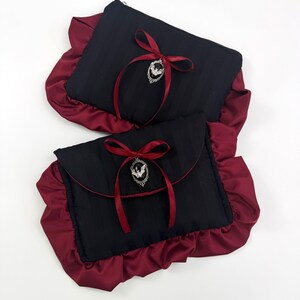 May include: Two black clutch bags with a burgundy satin ruffle trim. Each bag has a burgundy ribbon and a silver bat charm. One bag has a zipper closure, and the other has a flap closure.