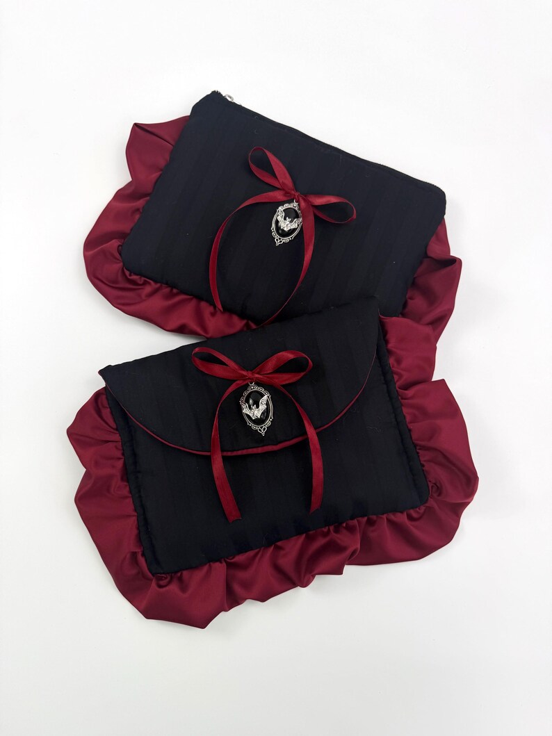 May include: Two black clutch bags with a burgundy satin ruffle trim. Each bag has a burgundy ribbon and a silver bat charm. One bag has a zipper closure, and the other has a flap closure.