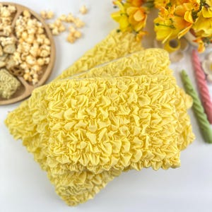 May include: Bright yellow textured fabric, possibly a cosmetic bag or pouch, is the focal point. The fabric has a ruffled, bubbly texture. A wooden tray with snacks and yellow flowers are in the background.