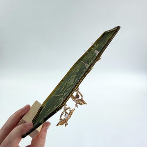 May include: A rectangular green fabric display board with a dark border, mounted on a light wooden stand. The board features a textured, patterned surface with several pairs of gold-toned moth-shaped earrings attached to the front.