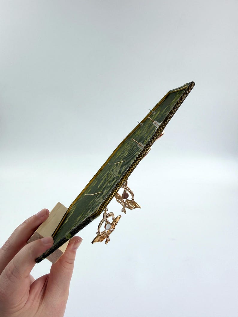 May include: A rectangular green fabric display board with a dark border, mounted on a light wooden stand. The board features a textured, patterned surface with several pairs of gold-toned moth-shaped earrings attached to the front.