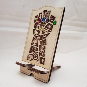 May include: Wooden phone stand shaped like a hand with the Infinity Gauntlet from the Marvel Cinematic Universe. The hand is holding the Infinity Stones, which are represented by colourful gems. The stand is engraved with the Marvel logo.