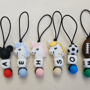 May include: Six colourful silicone baby teethers with different characters and letters. The teethers are attached to black cords. The characters include a unicorn, a football, a soccer ball, and Mickey Mouse. The letters are A, E, H, S, O, and M.