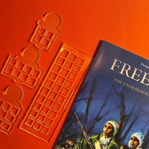May include: Three clear plastic stencils for a board game called "Freedom: The Underground Railroad". The stencils are shaped like houses with a grid of squares inside. The game title is visible on the blue cover of the game box.