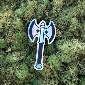 May include: A holographic sticker of a double-bladed axe. The axe features a black handle and a silver head with a rainbow sheen. Two stars and arrows are on the axe head. The sticker is set against a background of green moss.