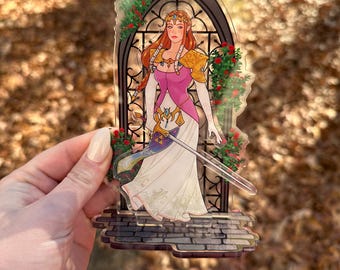 May include: A clear acrylic standee featuring a female character with long red hair, wearing a pink and white dress, and holding a sword. The character is set against a stained-glass window design with red roses.