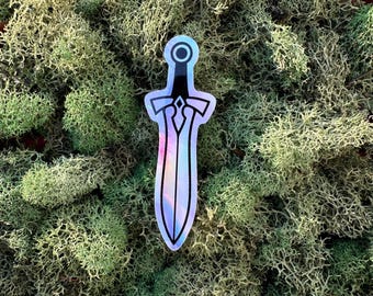 May include: A holographic sticker of a sword with a black handle and a silver blade. The sticker has a shiny, iridescent finish and is set against a backdrop of green moss.