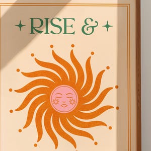 May include: A framed art print with the words "RISE &" in green above a stylized orange sun with a pink face. The sun has radiating lines and dots. The background is a light beige color.