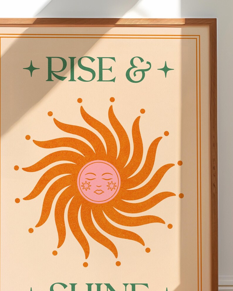 May include: A framed art print with the words "RISE &" in green above a stylized orange sun with a pink face. The sun has radiating lines and dots. The background is a light beige color.