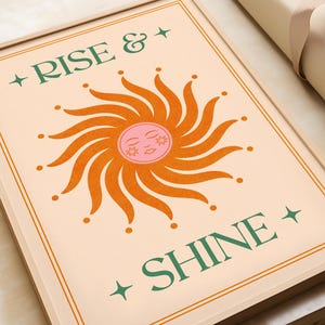 May include: A framed art print with the words "RISE & SHINE" in green lettering. The artwork features a stylized sun with orange rays and a pink face. The background is a light peach color, and the frame is a light wood tone.