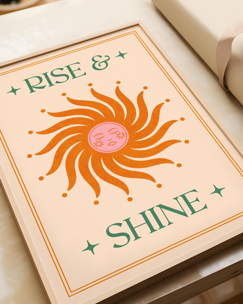 May include: A framed art print with the words "RISE & SHINE" in green lettering. The artwork features a stylized sun with orange rays and a pink face. The background is a light peach color, and the frame is a light wood tone.