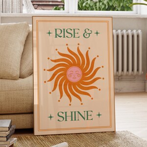May include: A framed art print with the words "RISE & SHINE" in green, with a stylized orange sun in the center. The sun has a pink face and is surrounded by orange rays. The print is in a wooden frame.